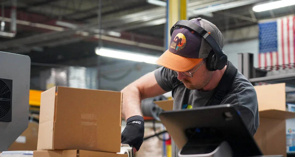 A 3D-Fuel production operator packing filament orders on the factory floor in Fargo, with an NDSU banner on the wall