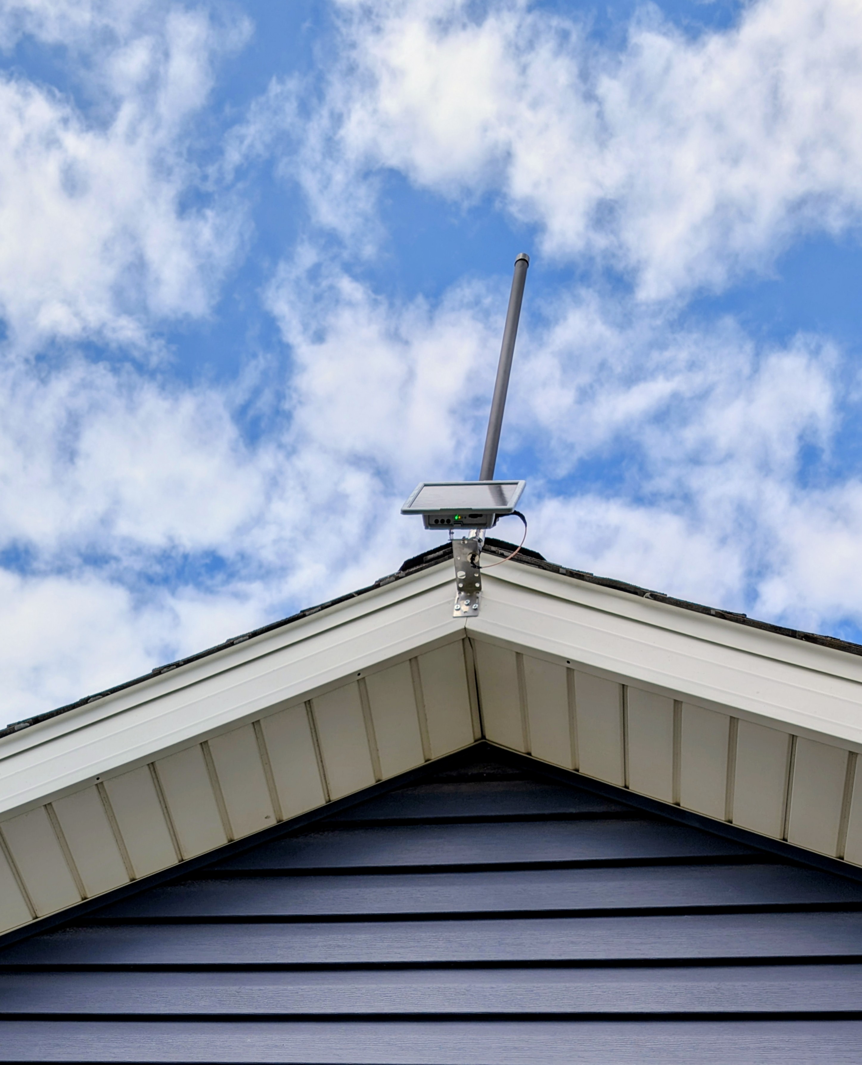 SenseCAP Solar Node P1-Pro mounted at the peak of a residential roof gable with a fiberglass LoRa antenna pointing into a partly cloudy sky