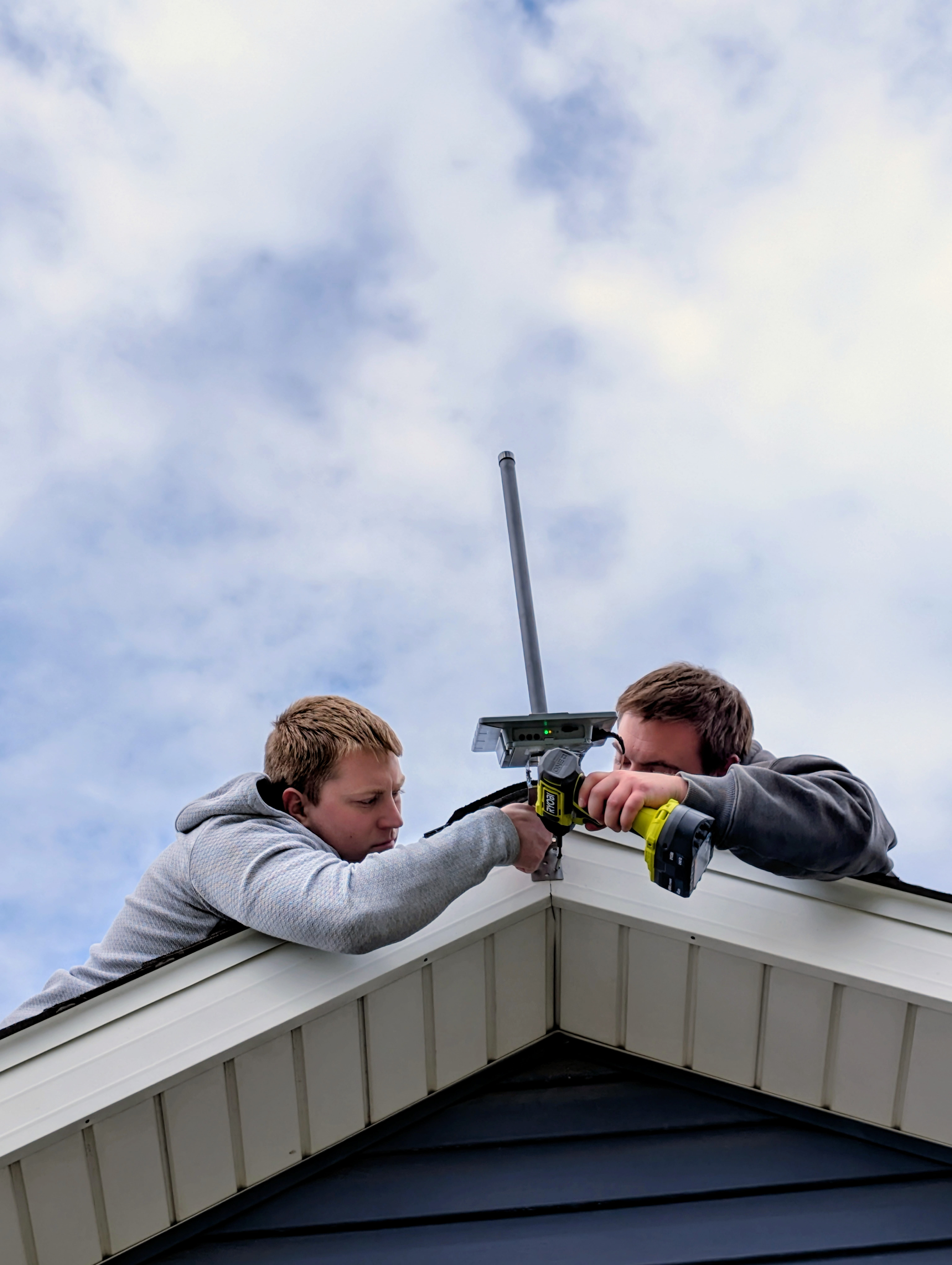 Two installers at a residential roof peak using a cordless drill to mount a Seeed SenseCAP Solar Node with a fiberglass LoRa antenna already attached